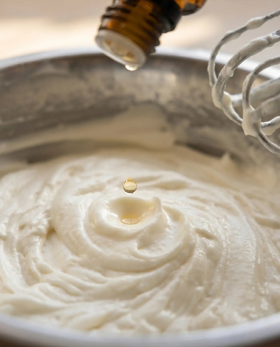 Single drop of essential oil meeting the surface of whipped grass-fed tallow in a stainless mixing bowl beside a stand mixer.