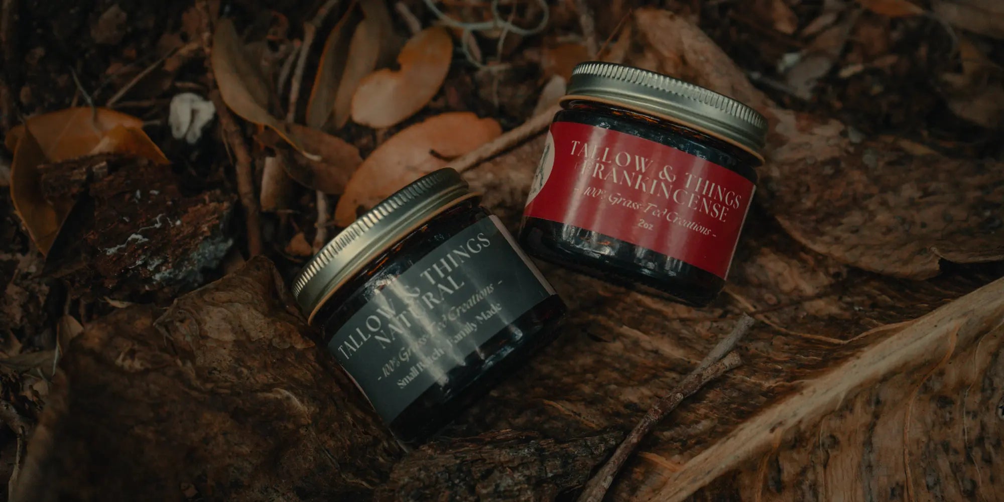 Two grass-fed tallow balm jars resting on a forest floor among fallen leaves