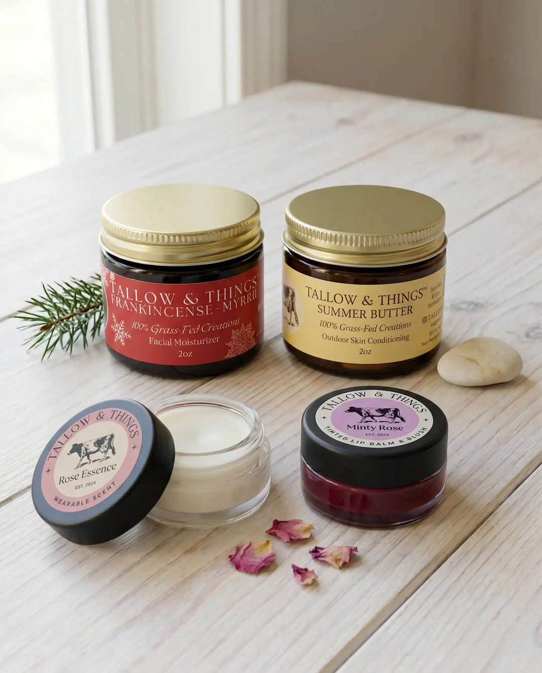Four Tallow & Things limited edition products — Frankincense & Myrrh Holiday Blend, Summer Butter, Rose Essence Wearable Scent, and Minty Rose Tinted Lip Balm — arranged together on a whitewashed wood surface with an evergreen sprig, dried rose petals, and a pale stone.