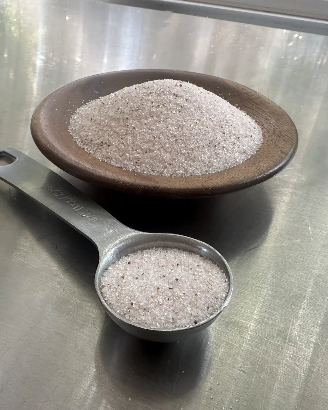 Pink-grey ancient sea salt in a wood dish and measuring spoon on steel.