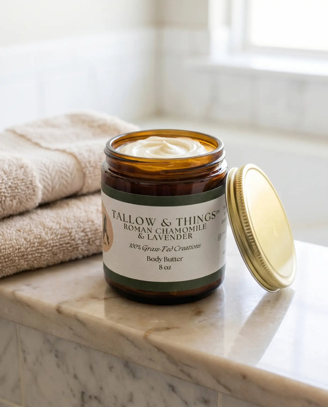 Open 8oz jar of Tallow & Things Roman chamomile and lavender body butter on a marble bath ledge with towels.