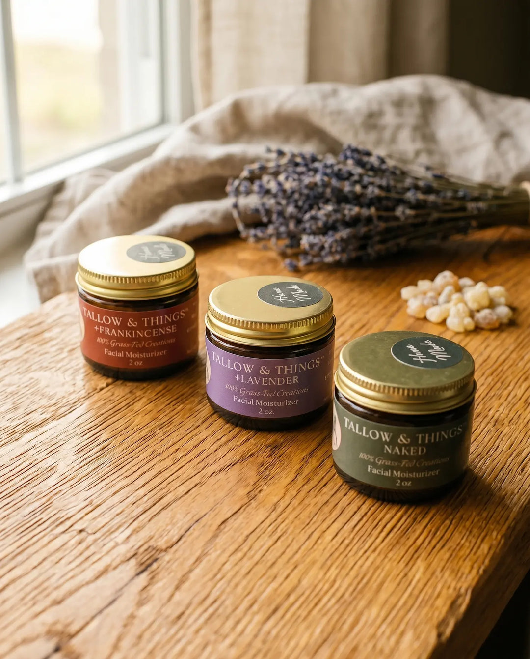 Three Tallow & Things facial moisturizer jars in frankincense, lavender, and naked on an oak table by a window.