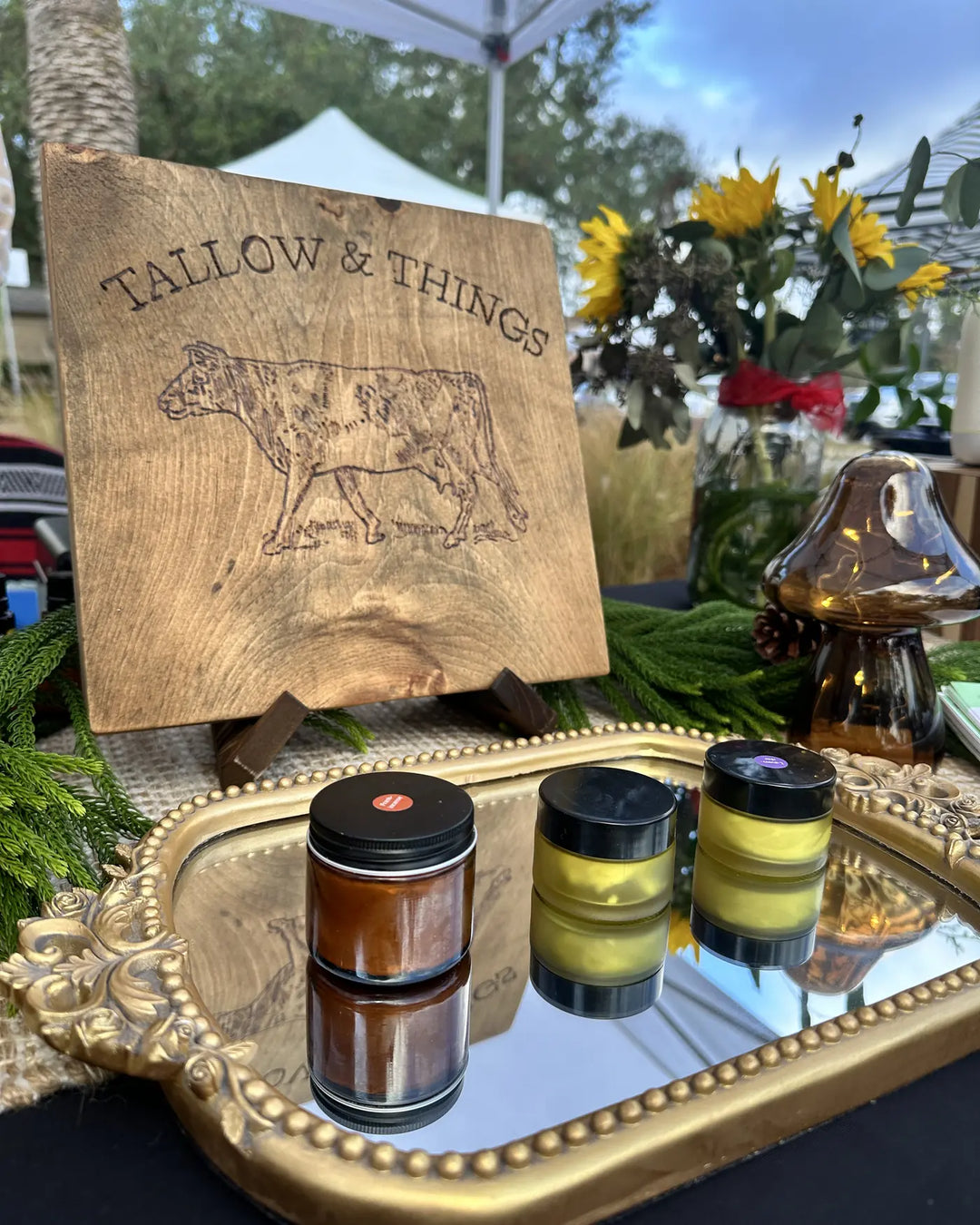Tallow and Things branded wood sign and product jars on a gold tray at a farmers market
