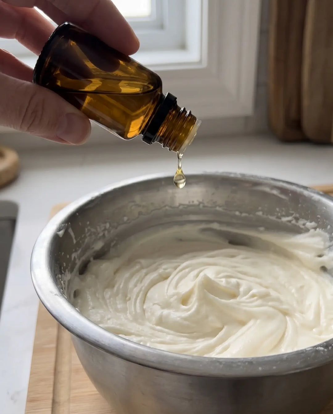 Unbranded amber essential oil bottle being tipped over a stainless mixing bowl of freshly whipped grass-fed tallow.
